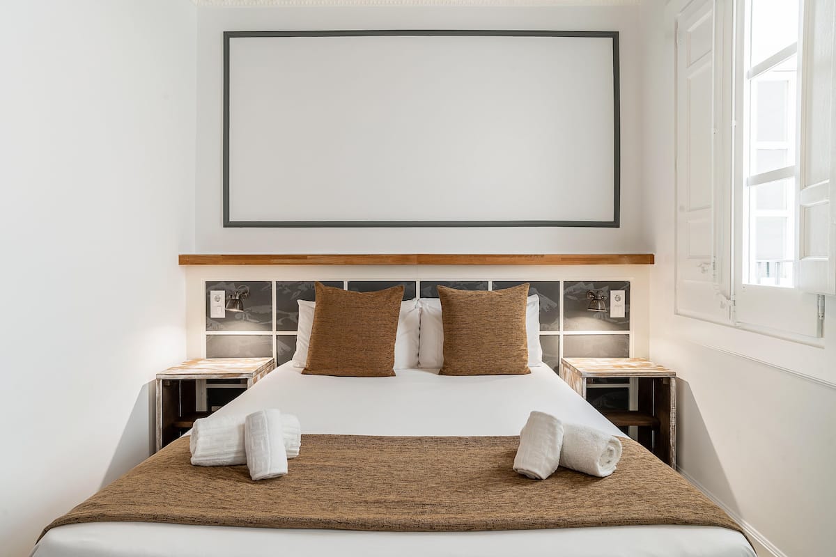 A neatly made bed with a brown throw blanket and two pillows, a bedside table with a lamp, and a large framed artwork above the bed.