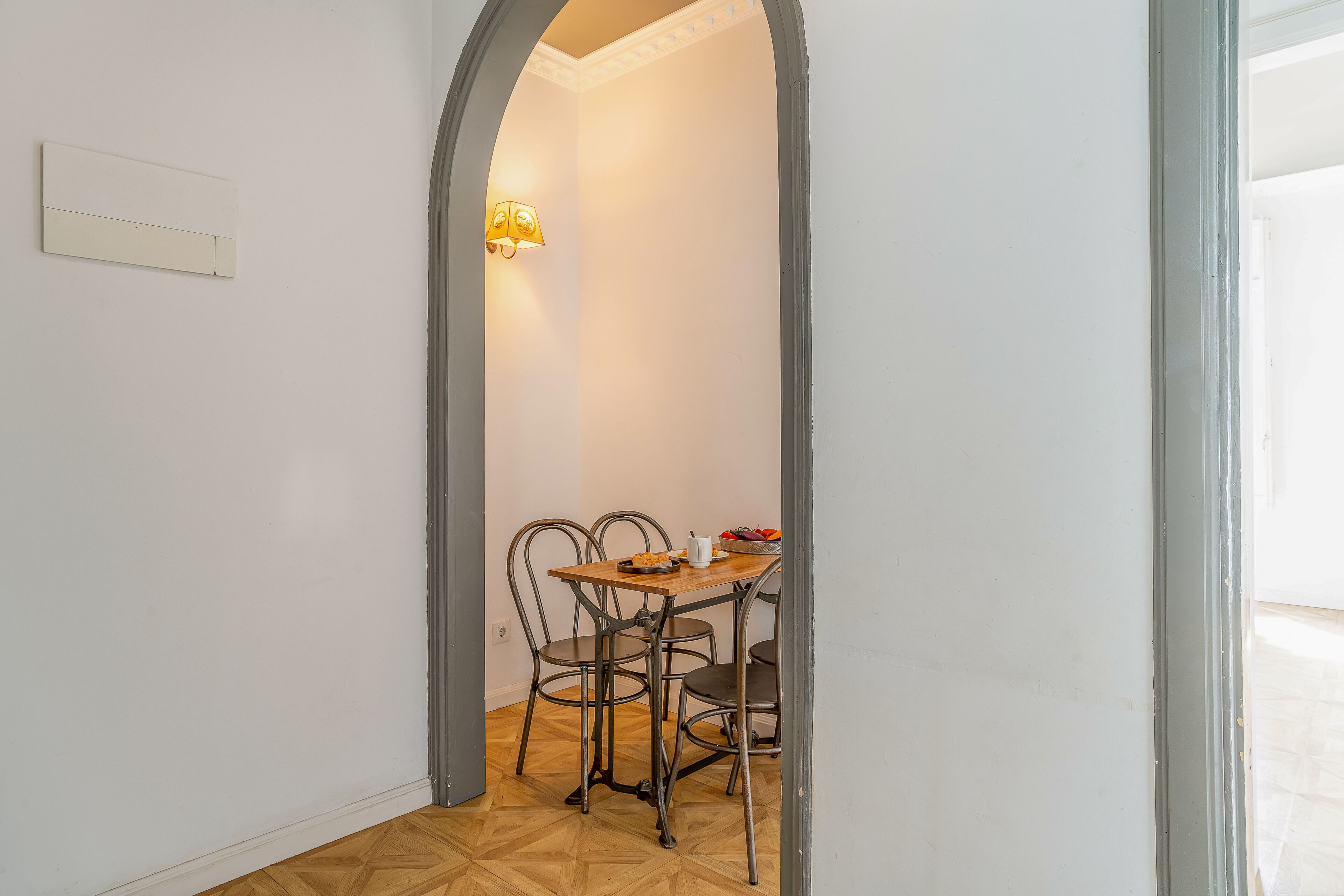 A dining area with a wooden table and four chairs, a light fixture on the wall, and a door leading to another room.