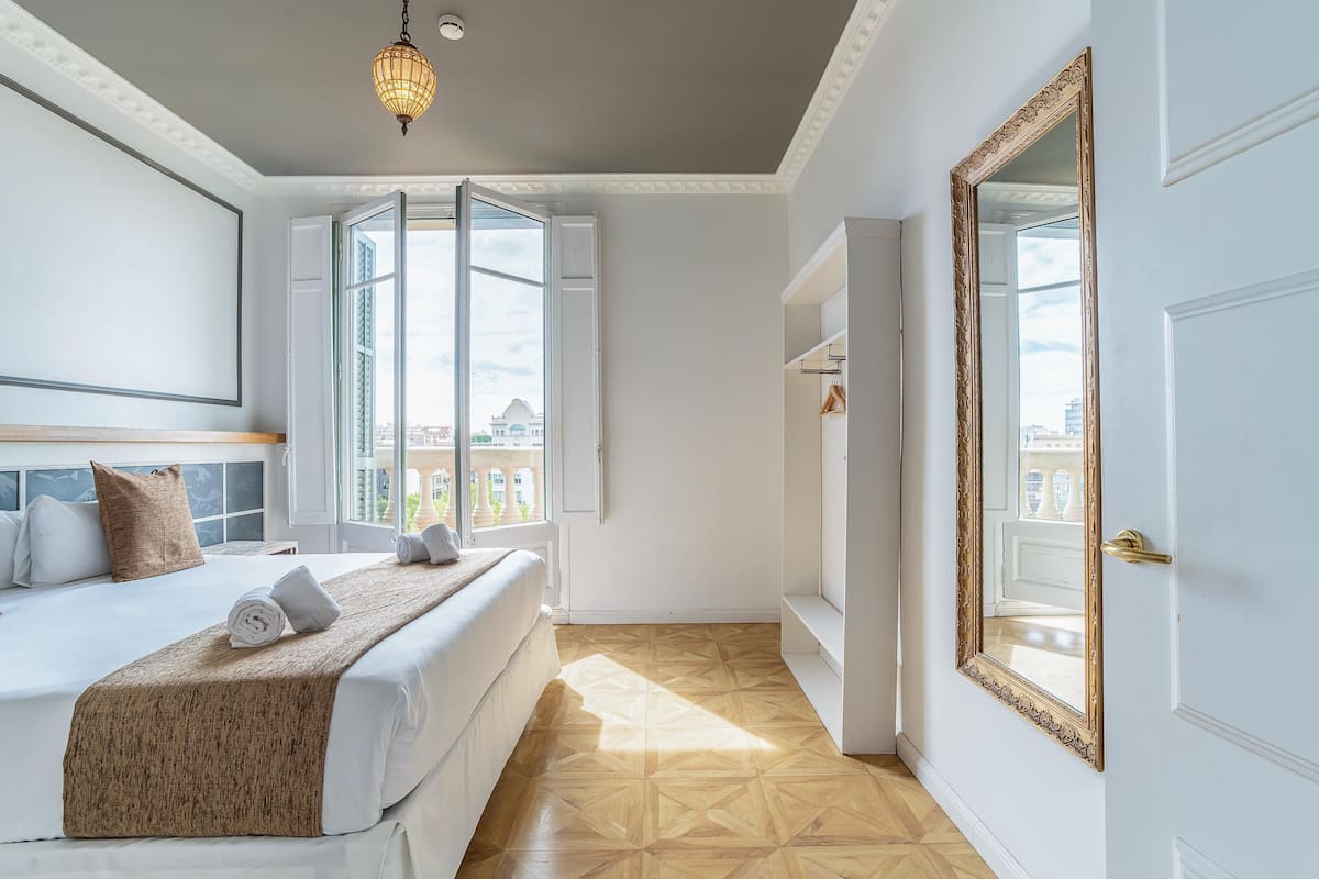 A hotel room with a bed, a large mirror, a window with shutters, and a view of buildings outside.