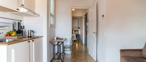 A narrow hallway with a kitchen on one side, a dining area with a table and chairs, and a living room with a sofa on the other side.