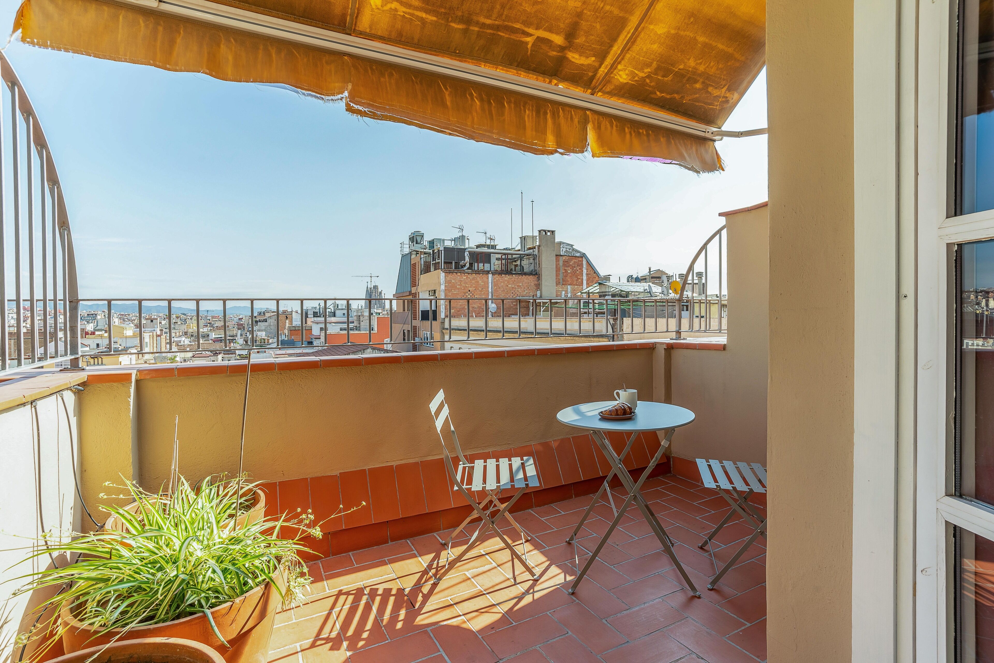 A balcony with a view of a cityscape, a potted plant, a table with a cup, and a chair.