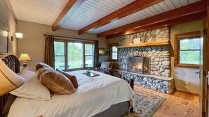 A bedroom with a stone fireplace, a large bed with pillows, and a view of the outdoors through the windows.