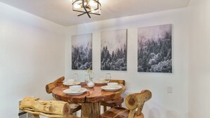 Zona pranzo con un tavolo rotondo in legno apparecchiato per quattro persone, circondato da sedie in legno con schienali originali. Tre quadri incorniciati raffiguranti paesaggi forestali adornano la parete sopra il tavolo.
