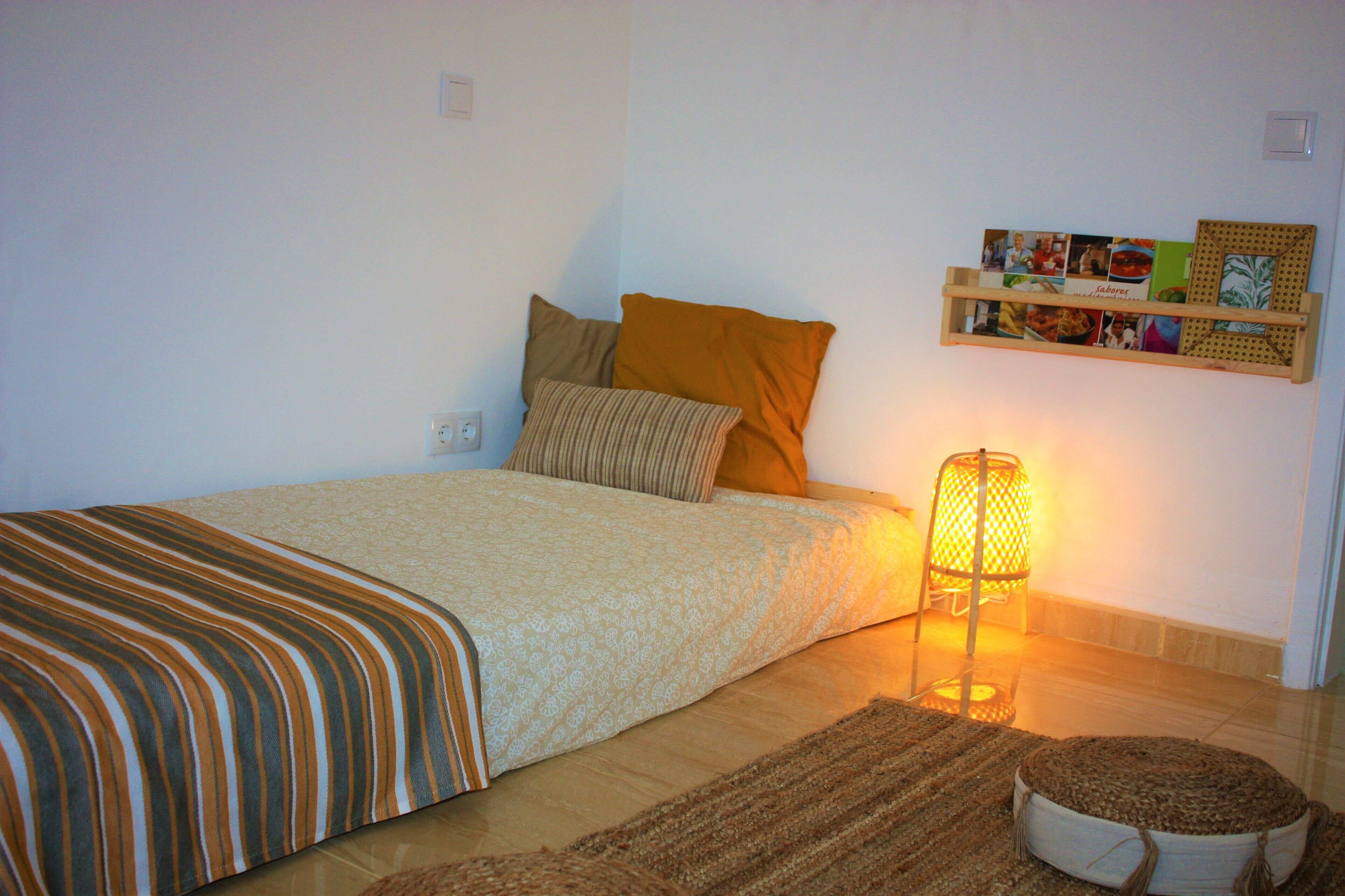 Un dormitorio ordenado con cama, almohadas, una lámpara de noche, un tapete y una estantería con fotos.