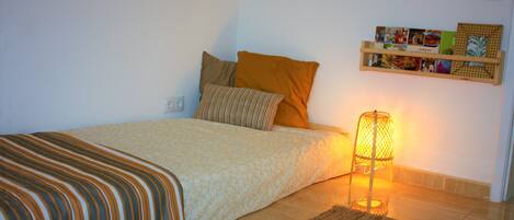 Un dormitorio ordenado con cama, almohadas, una lámpara de noche, un tapete y una estantería con fotos.