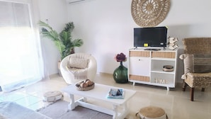 Una sala de estar con un sofá, una mesa de centro blanca, un mueble de televisión con televisor de pantalla plana, un adorno en la pared y una planta en maceta.