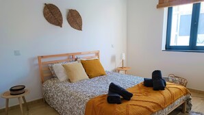 Un dormitorio ordenado con cama, mesitas de noche, una pequeña mesa redonda y una ventana con persianas.