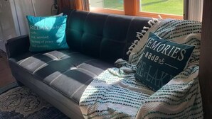 A cozy living room with a dark leather sofa, a patterned throw blanket, and two decorative pillows. - Cozy Cabin on Cobbossee Lake near Lakeside Marina (Winthrop)
