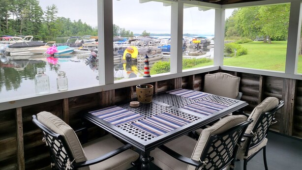 A covered outdoor seating area with a table and chairs, overlooking a marina with boats. - Cozy Cabin on Cobbossee Lake near Lakeside Marina (Winthrop)