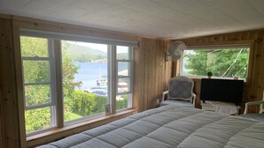 Un dormitorio con una cama grande, una silla, un televisor y un ventilador. Hay ventanas amplias que ofrecen vistas a un lago y árboles.
