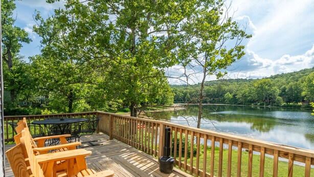 A wooden deck with Adirondack chairs and a table, overlooking a lake with trees and hills in the background. - Kiwanie Lodge, Large Lakefront Home, Sleeps 12 (Hardy)