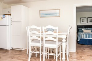 A dining area with a white table and chairs, a white refrigerator, and a bed with a blue bedspread in the background. - Beach Barn Lower - Lovely 3BR Lower Unit on the Oceanfront (Surf City)