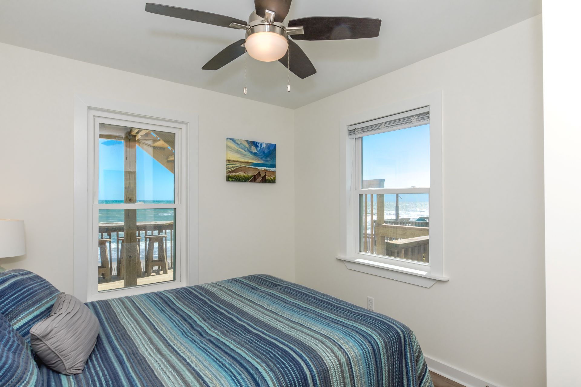 A bedroom with a bed, a ceiling fan, two windows with views of the ocean, and a painting on the wall.