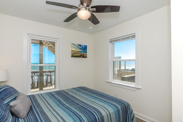 A bedroom with a bed, a ceiling fan, two windows with views of the ocean, and a painting on the wall.
