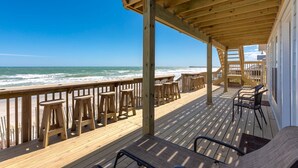 A wooden deck with a railing overlooking the ocean, featuring several wooden stools and a few chairs. - Beach Barn Lower - Lovely 3BR Lower Unit on the Oceanfront (Surf City)
