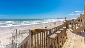 A wooden deck with a railing overlooking a beach with waves, a pier in the distance, and a clear blue sky. - Beach Barn Lower - Lovely 3BR Lower Unit on the Oceanfront (Surf City)