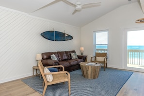 Quarto com sofá de couro marrom, mesa de centro redonda de madeira, cadeira de vime e tapete azul estampado. Um prancha de surf está fixada na parede, e há uma janela com persianas.