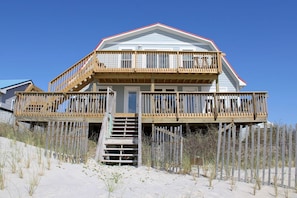 Una casa de playa de dos pisos con una terraza de madera y una escalera que lleva a la entrada.