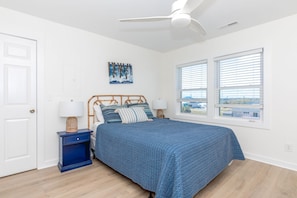 Quarto com cama, mesa de cabeceira, ventilador de teto, janela com persianas e uma obra de arte na parede.