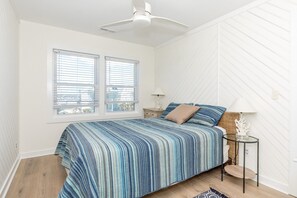 Une chambre à coucher avec un lit, deux fenêtres, un ventilateur de plafond, une table de chevet et une lampe.