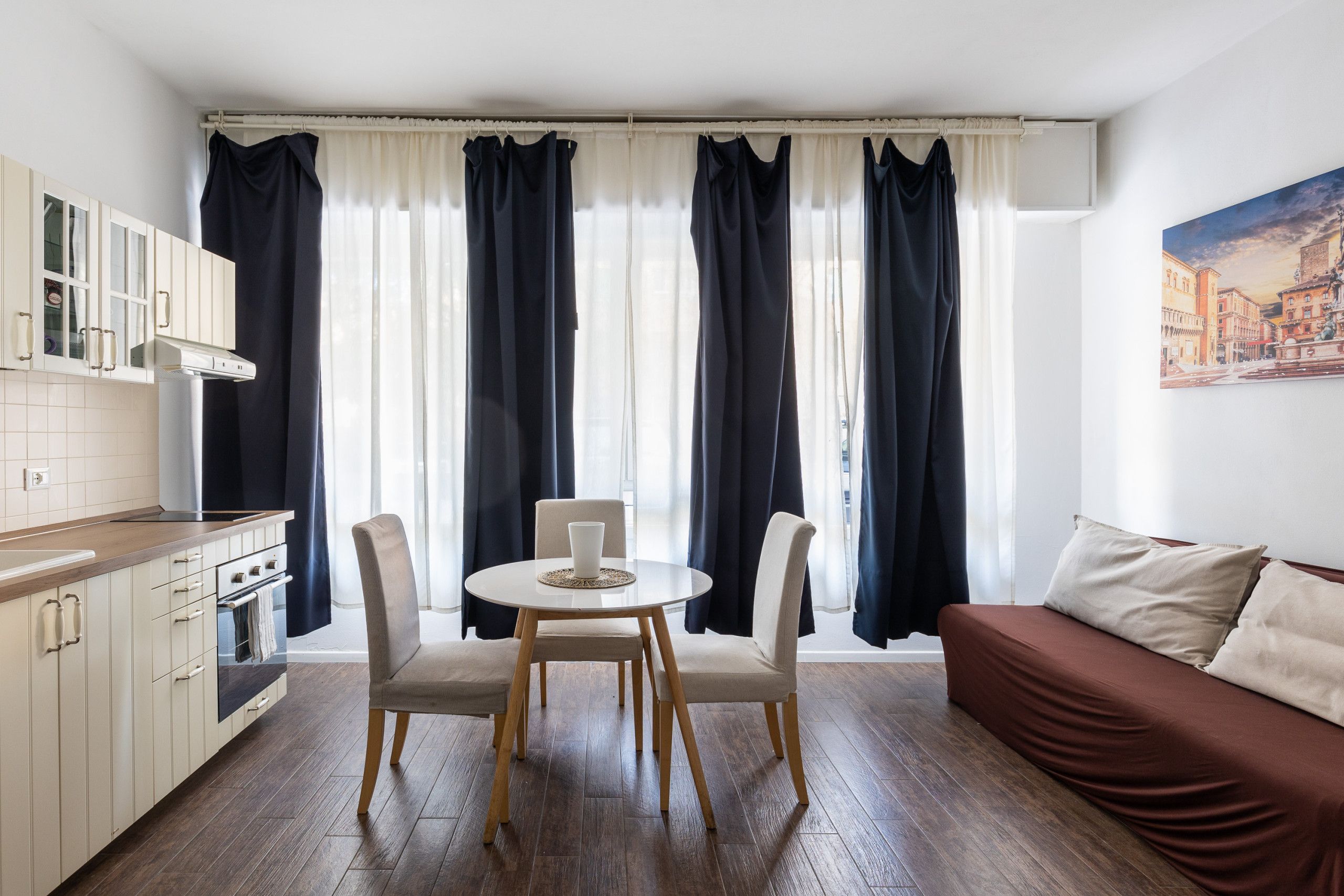Une cuisine moderne avec des armoires blanches et une hotte de cuisinière en acier inoxydable, un coin repas avec une table ronde et quatre chaises, et un salon avec un canapé marron et un tableau représentant un paysage urbain.