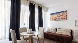 A room with a round table, two chairs, a sofa, and a painting of a cityscape on the wall.
