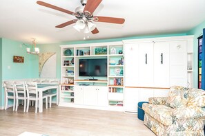 A living room with a ceiling fan, a television, a bookshelf, a dining area with chairs, and a patterned sofa. - @ Seas The Day - Charming 1BR Surf Condo; Community Pool (Surf City)