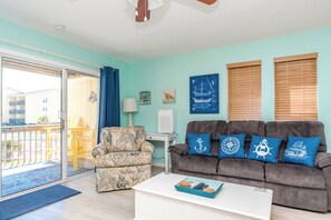 A living room with a sofa, armchair, and a coffee table with remote controls. - @ Seas The Day - Charming 1BR Surf Condo; Community Pool (Surf City)