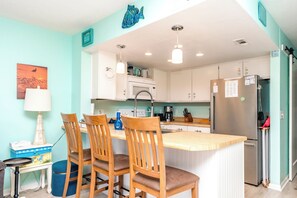 A kitchen with a central island, wooden chairs, a refrigerator, and white cabinets. - @ Seas The Day - Charming 1BR Surf Condo; Community Pool (Surf City)