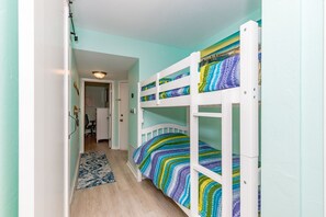 A bunk bed room with a patterned bedspread, a hallway leading to another room, and a door with a light fixture. - @ Seas The Day - Charming 1BR Surf Condo; Community Pool (Surf City)