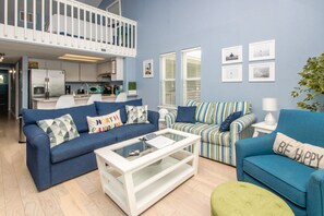 Un saló modern amb un sofà blau, un sofà amb ratlles i un otomà verd. Hi ha una taula de centre de vidre i una taula auxiliar blanca amb una làmpada. Es veu una cuina amb armaris blancs i un balcó al fons.