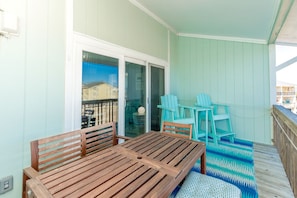 A balcony with a wooden table and chairs, a patterned rug, and a view of buildings through a glass door. - Sun And Surf - Top Floor 2 BR Surf Condo - Community Pool (Surf City)