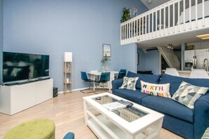 A modern living room with a flat-screen TV, a blue sofa with patterned cushions, a glass coffee table, and a dining area with a white table and blue chairs. - Sun And Surf - Top Floor 2 BR Surf Condo - Community Pool (Surf City)