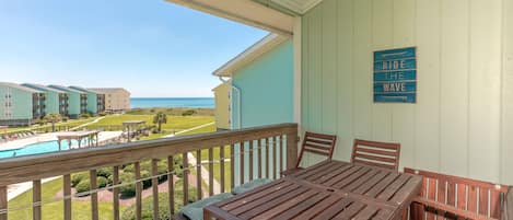 A balcony with a view of a swimming pool and ocean, wooden furniture, and a sign that says 'RIDE THE WAVE'.