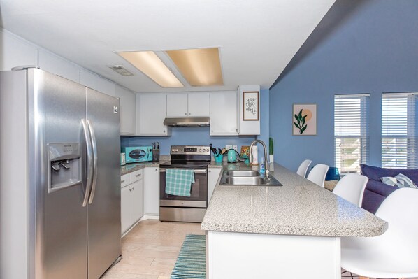 A modern kitchen with stainless steel appliances, a granite countertop, and a dining area with white chairs. - Sun And Surf - Top Floor 2 BR Surf Condo - Community Pool (Surf City)