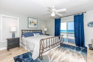 Una camera da letto con un letto, un comodino, una lampada, una ventola a soffitto e vista sull'oceano dalla finestra.