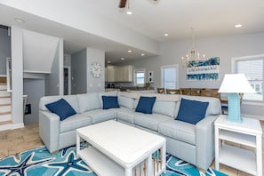 A modern living room with a sectional sofa, a coffee table, and a dining area with a chandelier. - Borders The Beach - 5BR Oceanfront Home with Amazing Views! (Surf City)