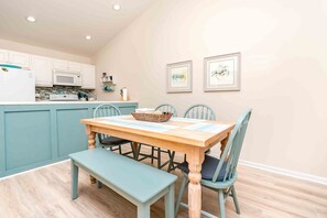 Uma área de refeições com mesa de madeira, quatro cadeiras, um banco e uma cozinha ao fundo.