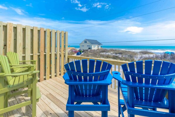 A wooden deck with two blue Adirondack chairs and a green chair, overlooking a beach with a house in the background.