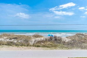 Vista da praia com dunas de areia, céu claro e um guarda-sol azul ao longe.