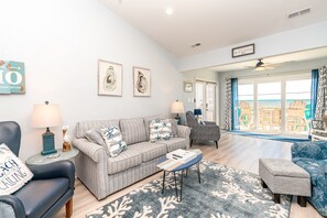 A living room with a sofa, armchairs, a coffee table, and a view of the beach through large windows.