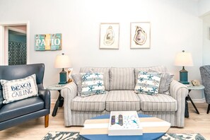 Una sala de estar con un sofá a rayas, un sillón negro y una mesa de centro. Hay dos lámparas sobre mesas auxiliares, un cuadro enmarcado sobre el sofá y un libro en la mesa de centro.