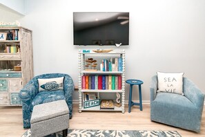 Un soggiorno con una libreria, una poltrona blu, un poggiapiedi grigio, uno sgabello e una TV fissata al muro.