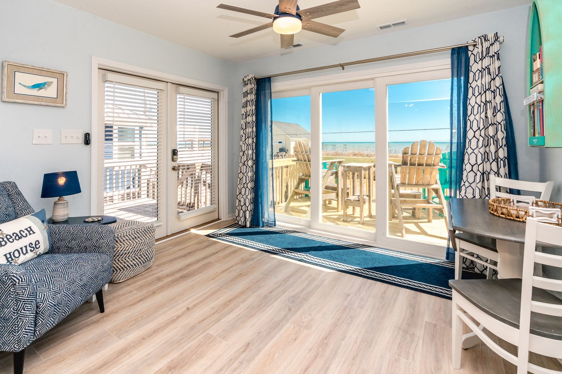 Ein Zimmer mit Holzboden, Deckenventilator, einer Couch mit gemusterten Kissen, einem Tisch mit einem gewebten Korb und Blick auf den Strand durch große Glasüren.