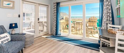 Quarto com piso de madeira, ventilador de teto, sofá com almofadas estampadas, mesa com cesta de vime e vista para a praia através de grandes portas de vidro.