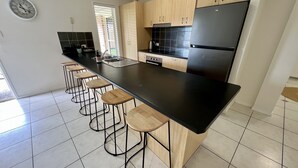 Une cuisine moderne avec un plan de travail noir, des tabourets en bois, un réfrigérateur noir et une fenêtre équipée de stores.