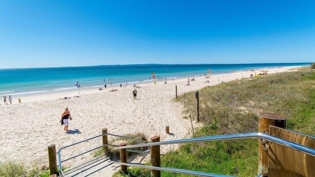 Une plage avec un escalier en bois qui descend jusqu'au sable, un ciel bleu clair et une mer calme.
