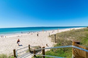 Una playa con una escalera de madera que baja a la arena, un cielo azul despejado y un mar tranquilo.