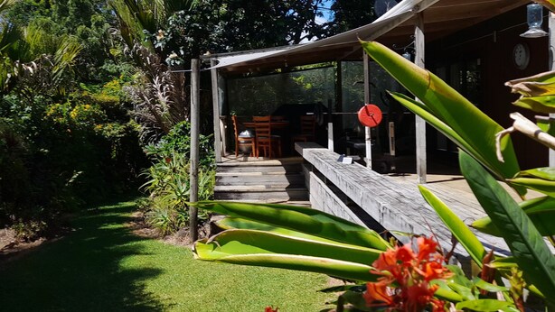 Un área de estar al aire libre con techo, con un banco y sillas de madera, rodeada de abundante follaje y un pequeño jardín.
