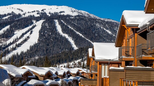 Et fjeldhotel med flere træhytter, en skibakke i baggrunden og en klar blå himmel.